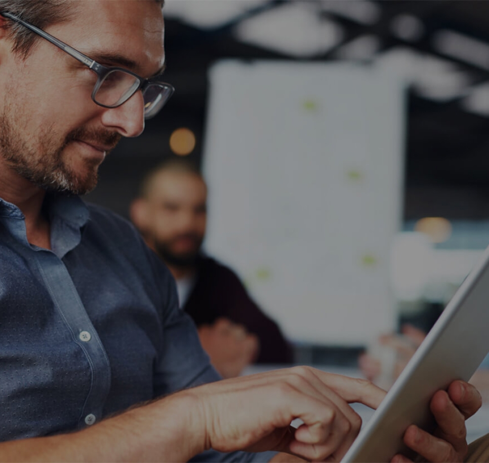 A man with a beard and glasses working on an ipad with a conference room of people behind him.