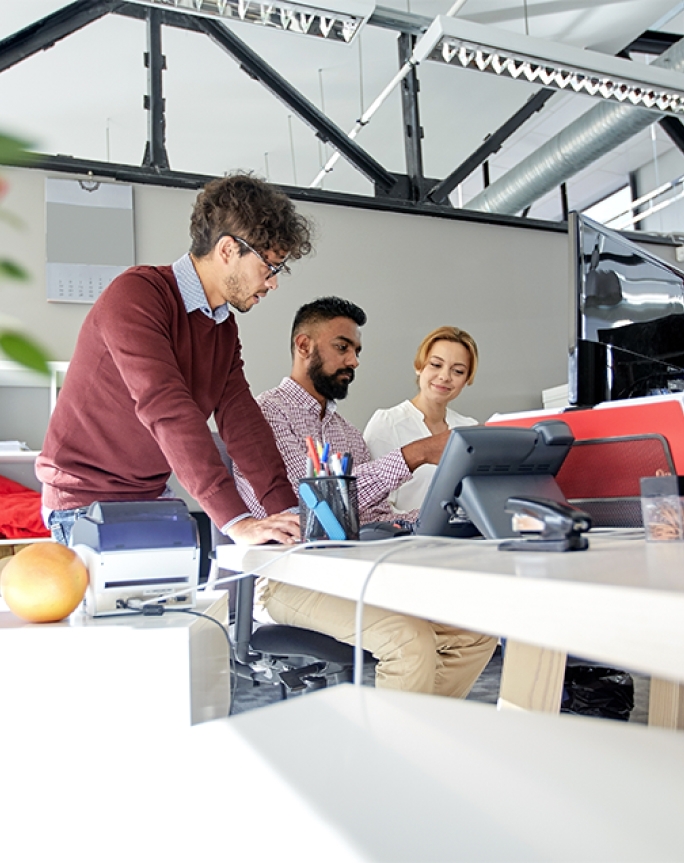 A group of people standing around working at an office desk in a large open office.