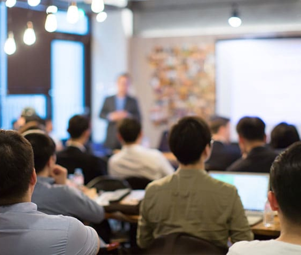 A faded image of a person teaching a conference room of students.