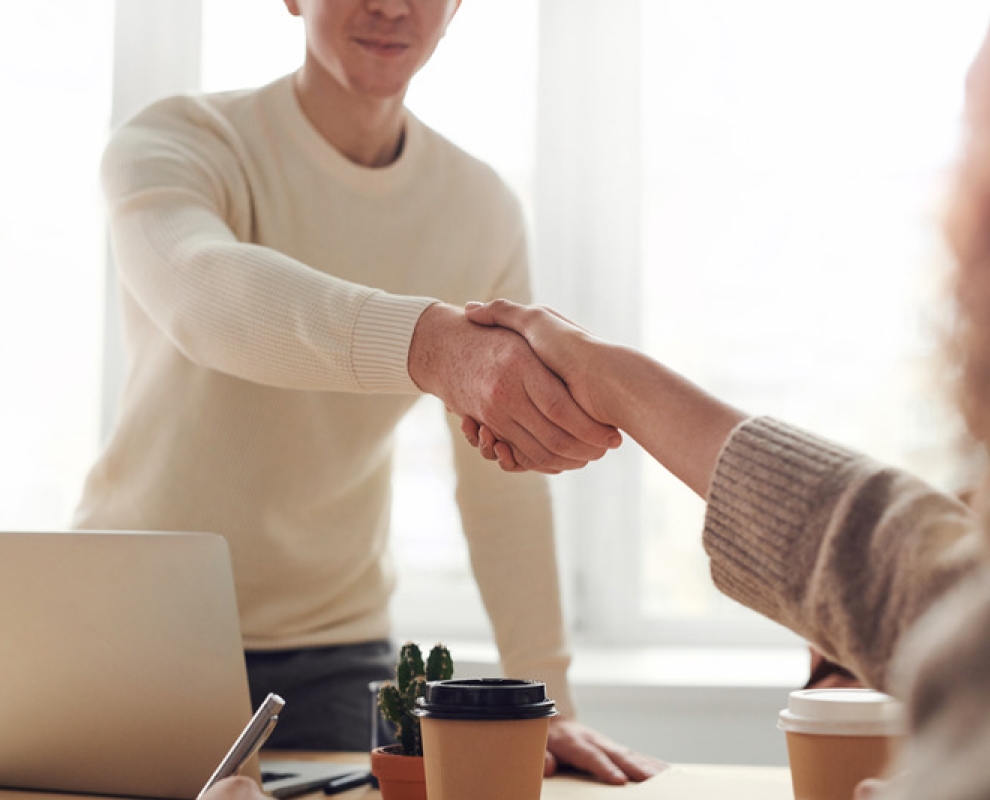 Two people shaking hands over a desk with a cactus and cup of coffee.